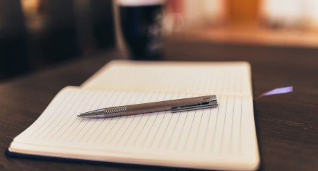 A notepad with a pen on top of it, and a black coffee cup in the background.